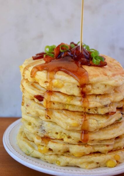 Tall stack of Sweet Corn Pancakes with Bacon topped with sliced scallions and drizzled with maple syrup