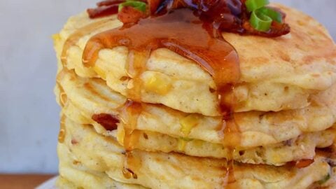 Tall stack of Sweet Corn Pancakes with Bacon topped with sliced scallions and drizzled with maple syrup