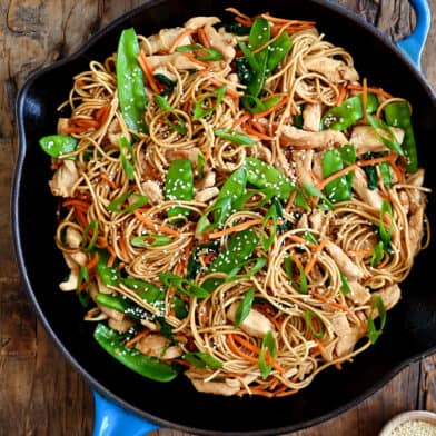 A large skillet containing stir-fried noodles, chicken pieces, snow peas, spinach and shredded carrots.