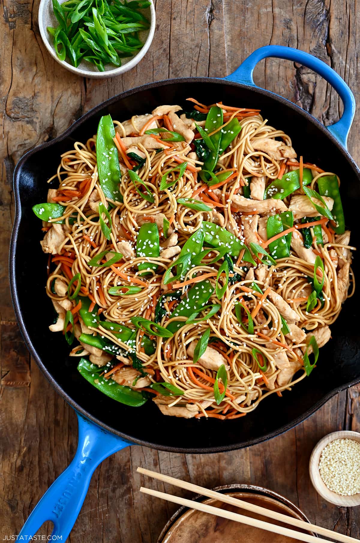 A large skillet containing stir-fried noodles, chicken pieces, snow peas, spinach and shredded carrots.