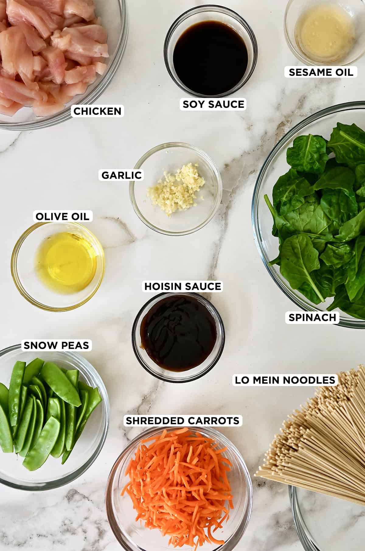 Various sizes of clear bowls containing olive oil, spinach, lo mein noodles, shredded carrots, snow peas, hoisin sauce, minced garlic, sesame oil, soy sauce and raw chicken pieces.