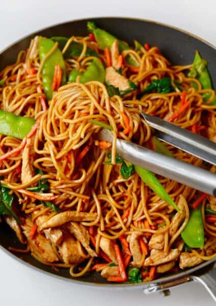 A tongs grabs chicken lo mein with snap peas and carrots from a wok.