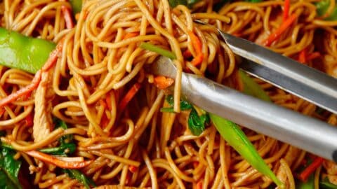 A tongs grabs chicken lo mein with snap peas and carrots from a wok.