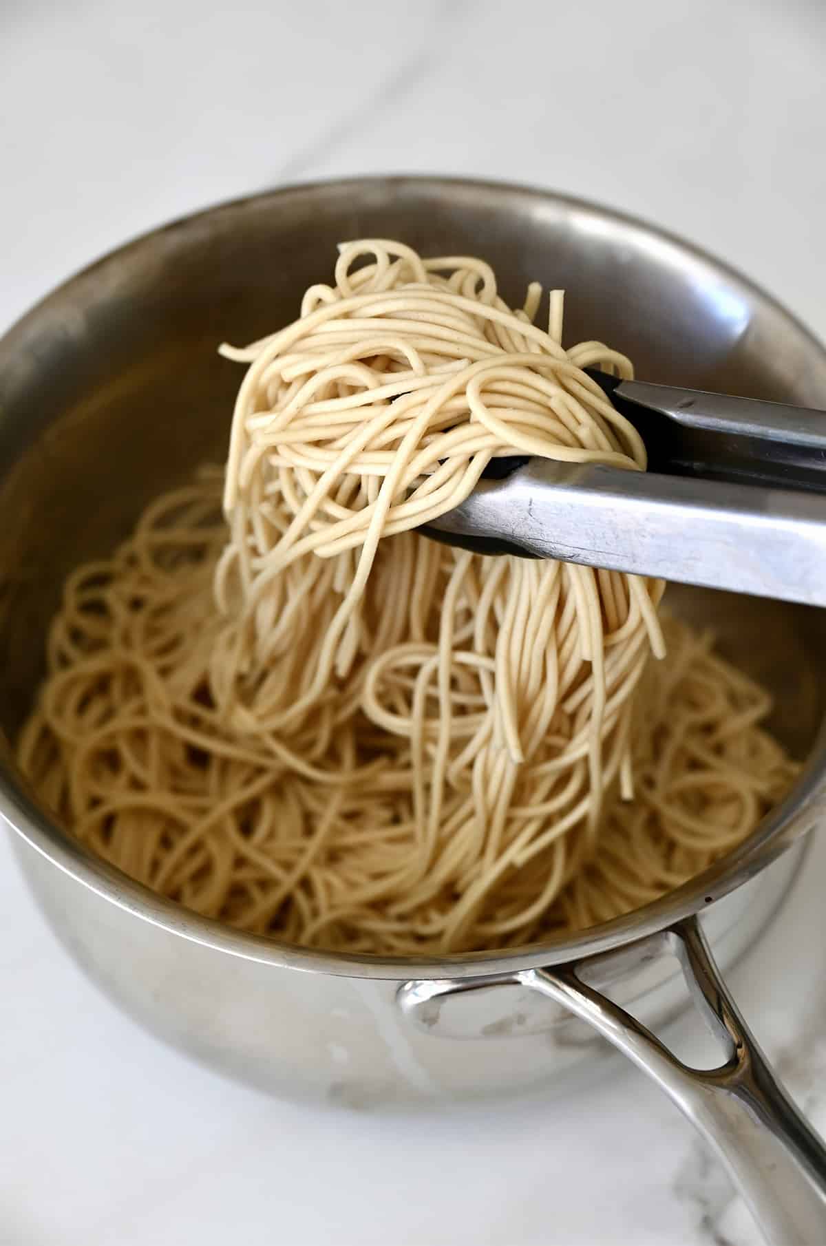 Tongs pulling at cooked lo mein noodles in a pan.