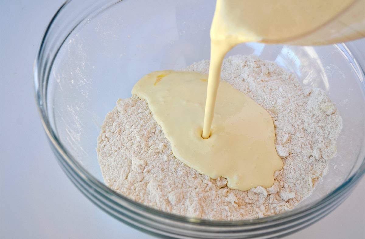A measuring cup pouring wet ingredients into dry ingredients that are in a large glass mixing bowl.