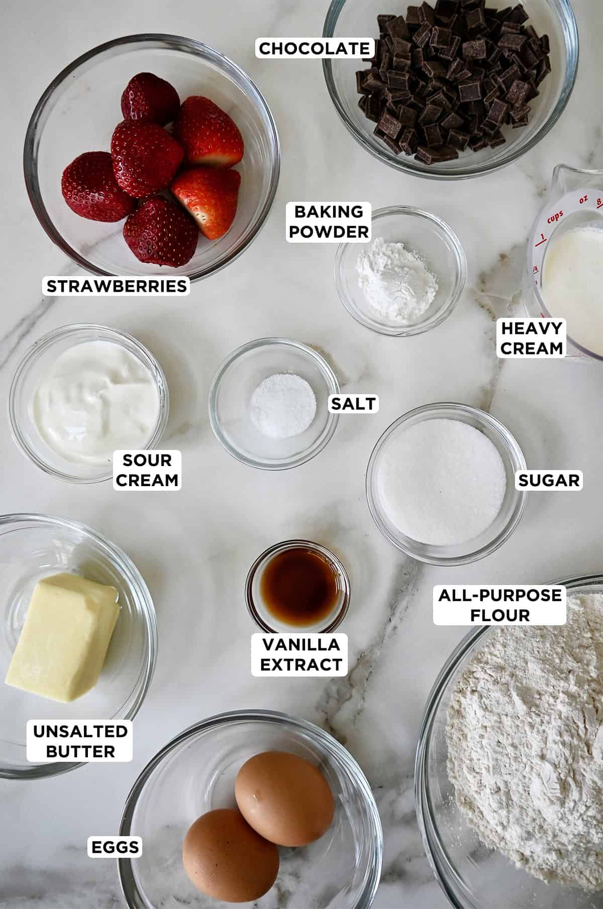 Glass bowls containing ingredients for strawberry scones, including fresh strawberries, sour cream, butter, eggs, flour, vanilla extract, sugar, salt, baking powder, heavy cream and chocolate chips.