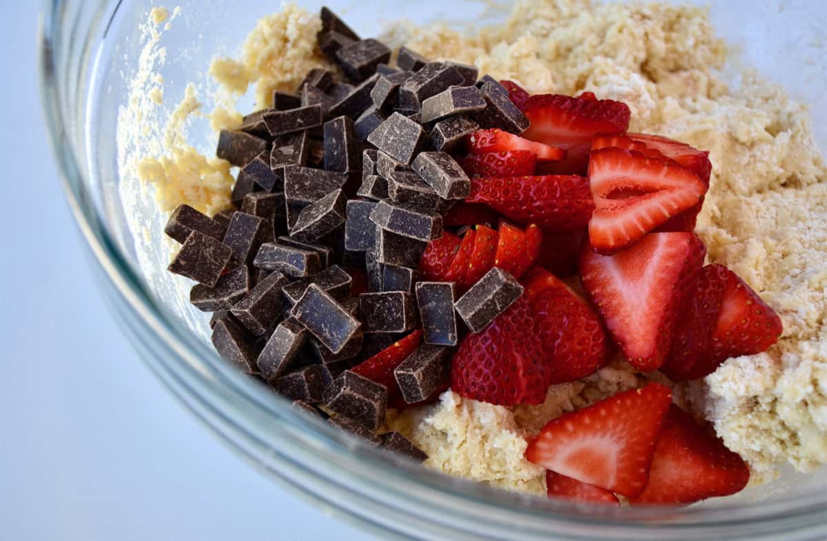 Strawberry slices and chocolate chunks atop scone batter in a glass bowl.