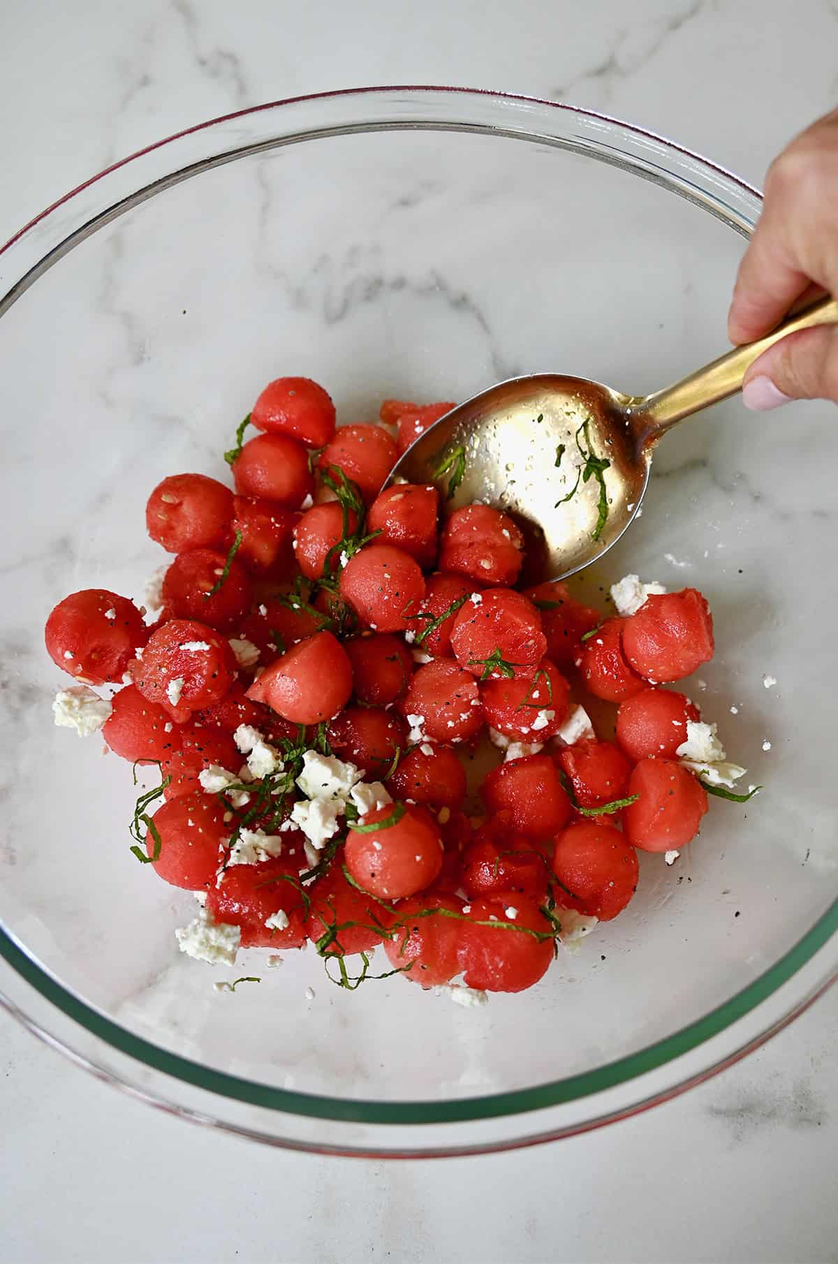 Watermelon balls with crumbled feta and fresh mint being tossed with a tangy lime dressing in a large bowl.