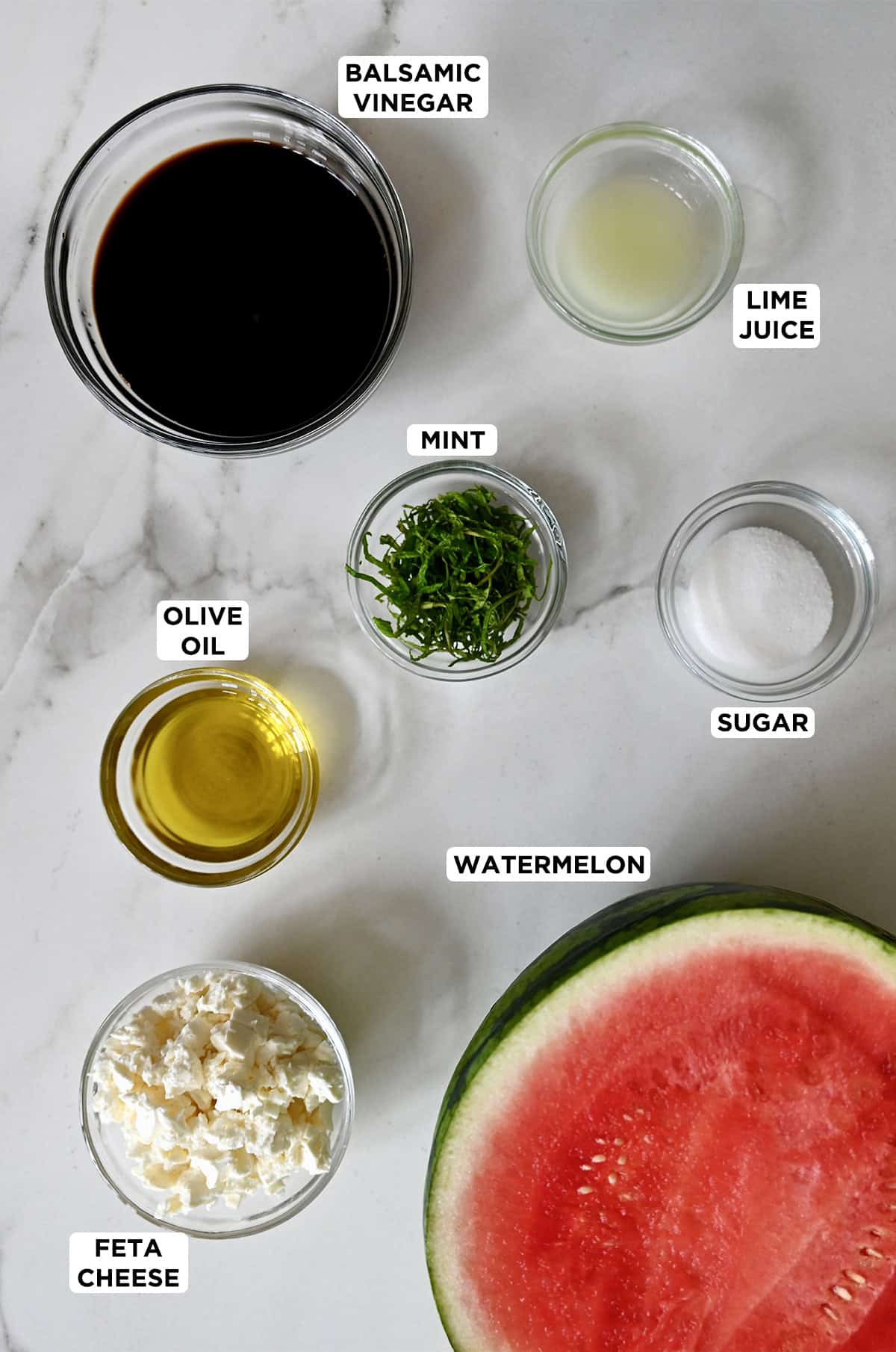 A large watermelon sliced in half next to small bowls containing crumbled feta cheese, olive oil, mint, sugar, lime juice and balsamic syrup.