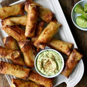 A top down view of baked egg rolls with small bowls of avocado dip, tajin and lime