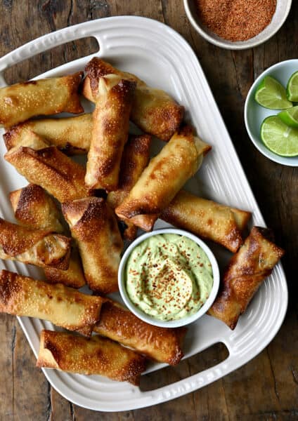 A top down view of baked egg rolls with small bowls of avocado dip, tajin and lime