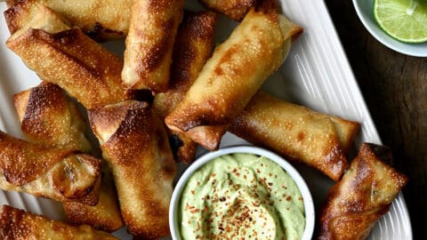 A top down view of baked egg rolls with small bowls of avocado dip, tajin and lime