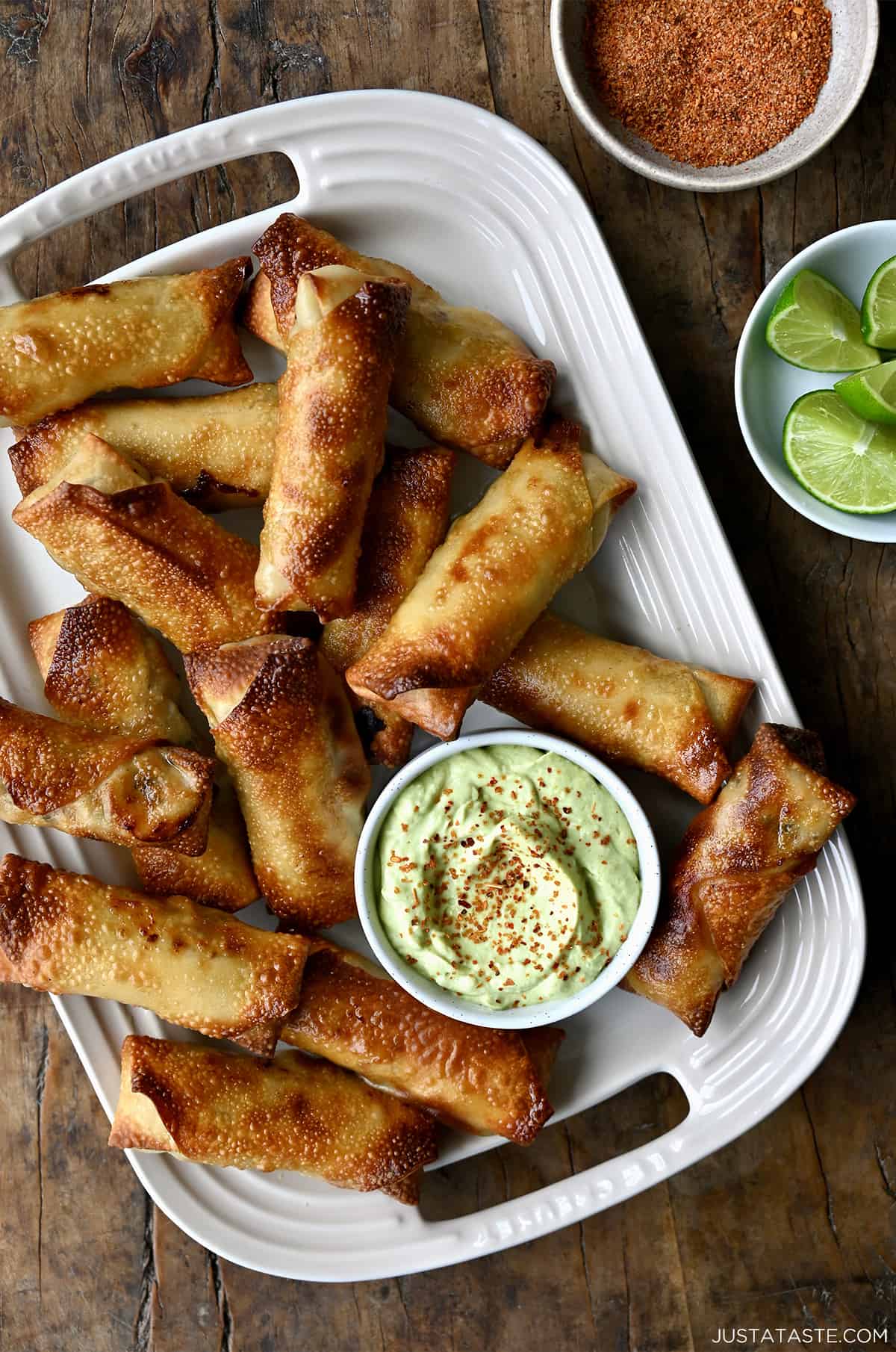 A top down view of baked egg rolls with small bowls of avocado dip, tajin and lime
