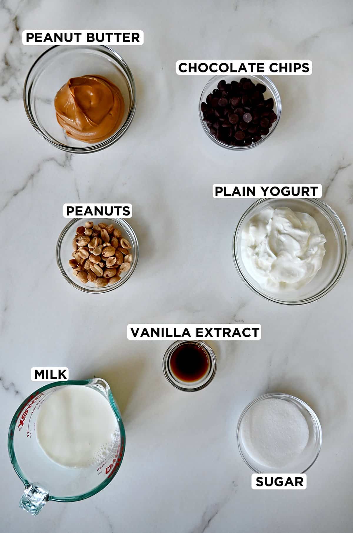 A top-down view of various sizes of clear bowls plus a liquid measuring cup containing peanut butter froyo ingredients, including creamy peanut butter, chocolate chips, plain yogurt, vanilla extract, sugar, peanuts and milk.