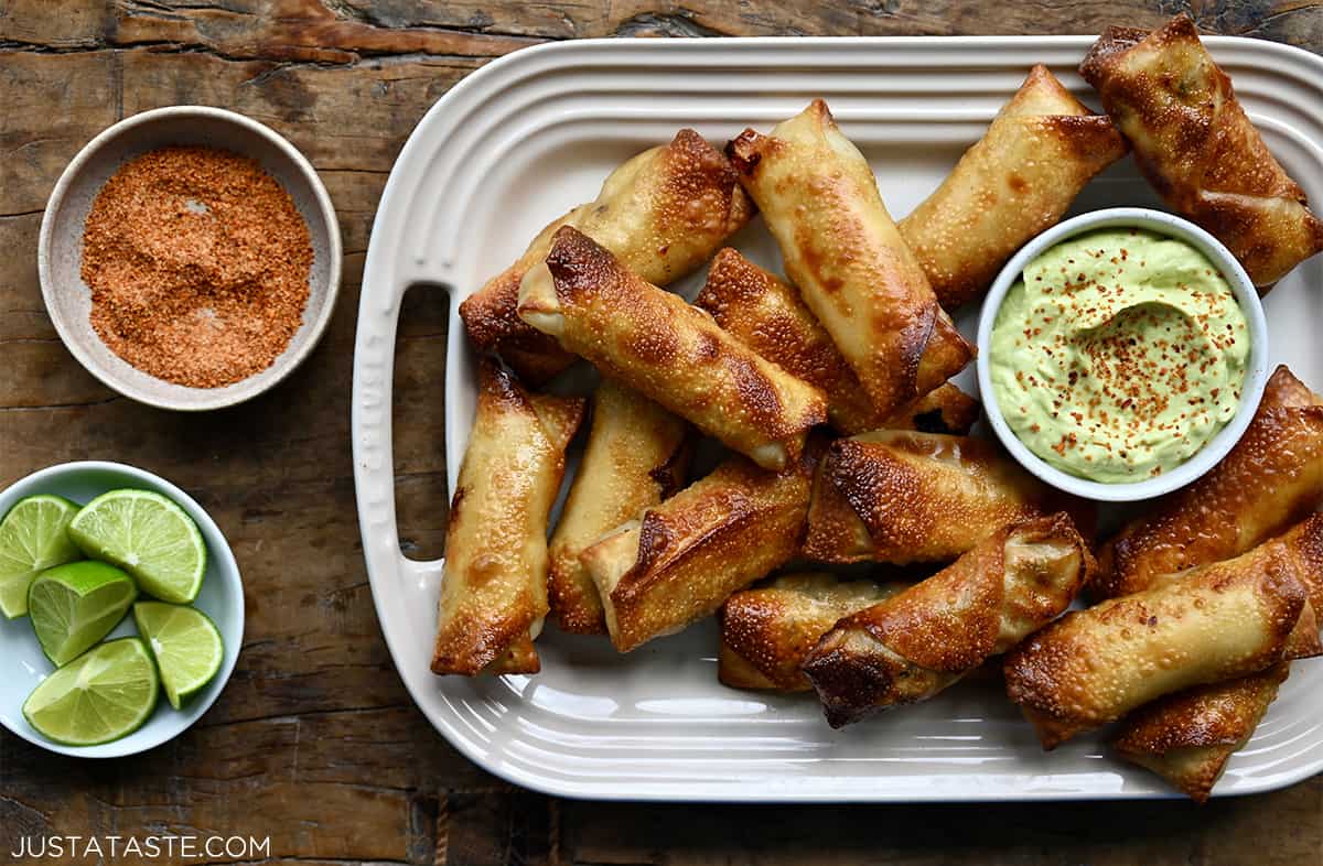 A top-down view of egg rolls on a white platter with bowls of avocado dip, takin and limes