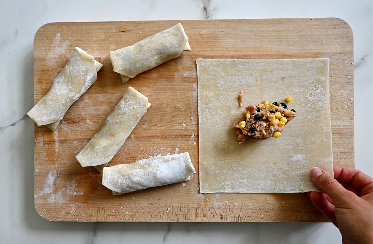 A cutting board topped with egg roll wrappers being filled and rolled