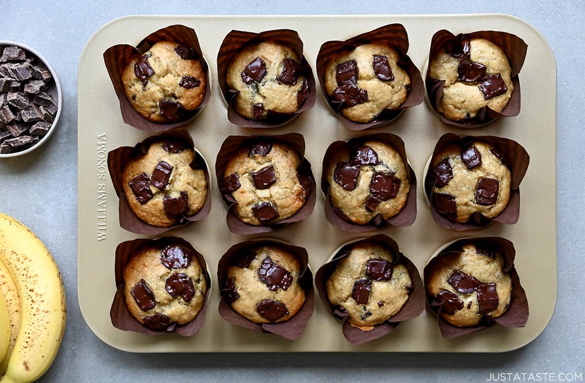 Banana chocolate chip muffins in a muffin pan next to a small bowl filled with chocolate chips.