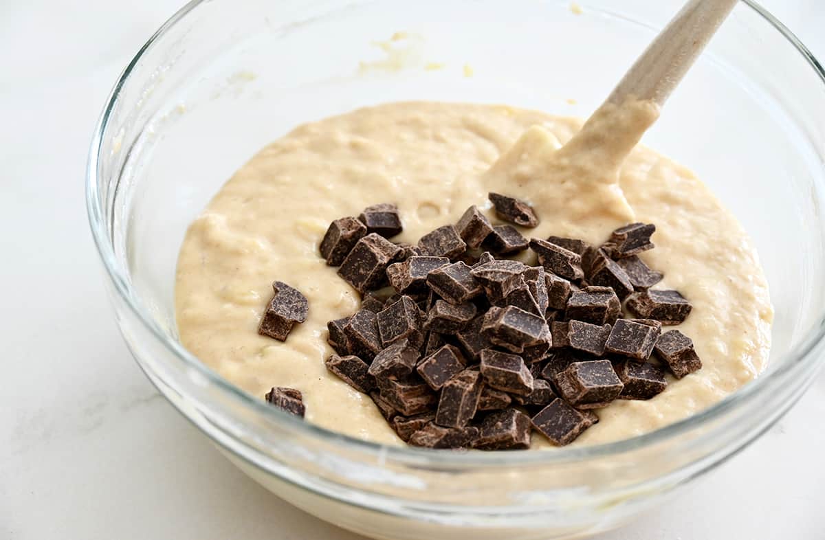 Chocolate chunks atop banana bread batter in a glass bowl with a spatula.