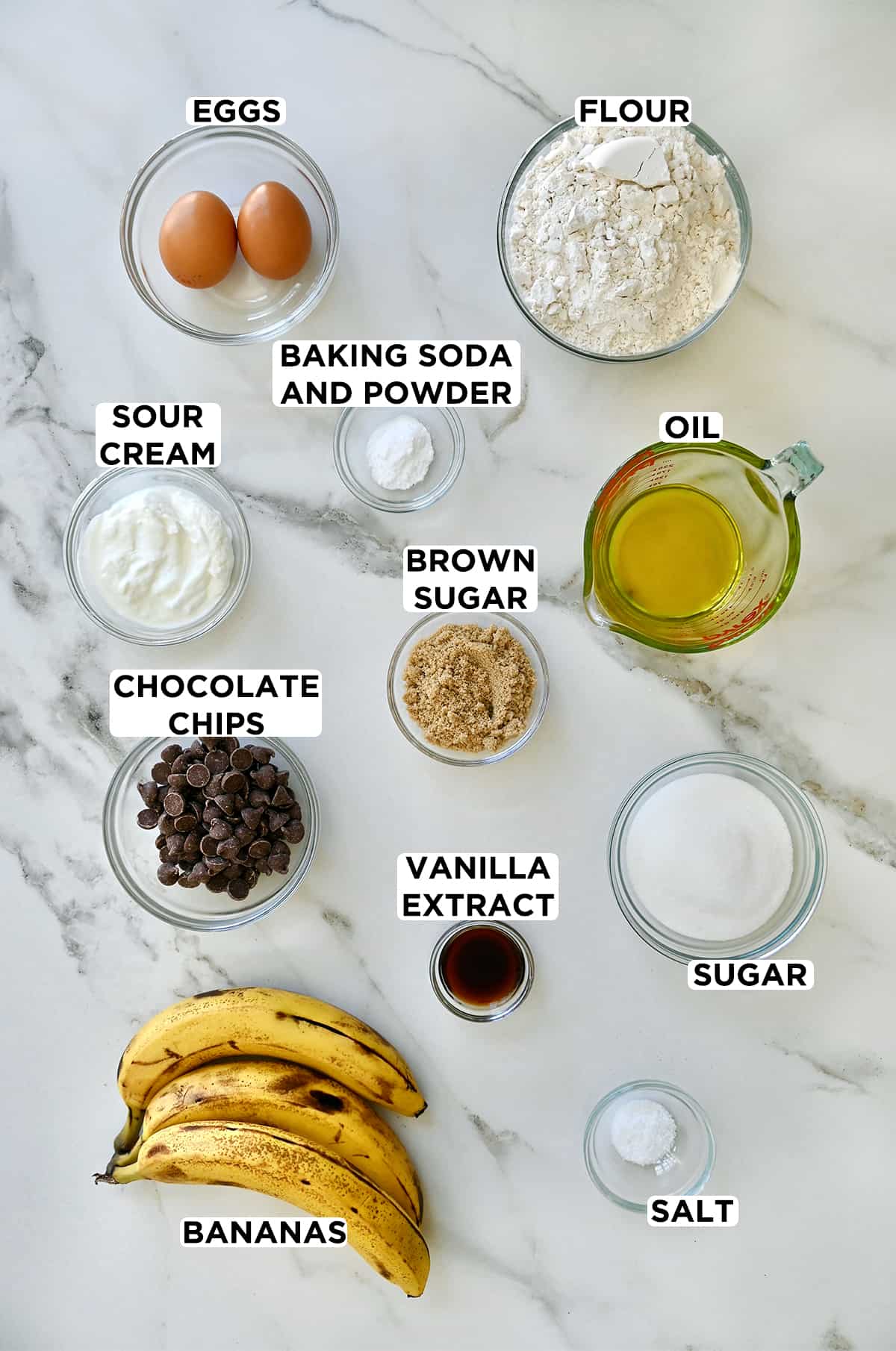 Three ripe bananas next to various sizes of glass bowls containing eggs, baking soda and powder, flour, sour cream, chocolate chips, vanilla extract, sugar, salt and a measuring cup filled with olive oil.