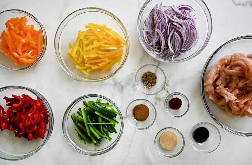 A top-down view of various sizes of clear bowls containing sliced bell peppers, sliced red onions, sliced chicken breasts and seasonings