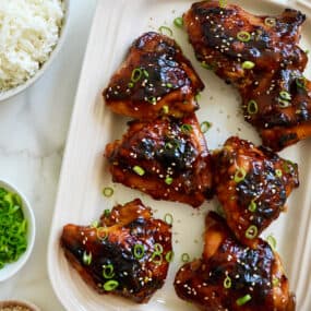 A top-down view of Glazed Honey Soy Chicken Thighs on a white serving platter next to bowls containing white rice, sliced scallions and sesame seeds