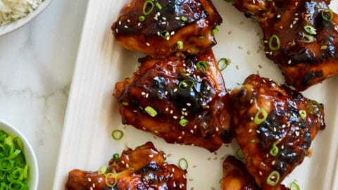 A top-down view of Glazed Honey Soy Chicken Thighs on a white serving platter next to bowls containing white rice, sliced scallions and sesame seeds