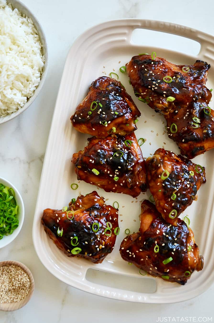 A top-down view of Glazed Honey Soy Chicken Thighs on a white serving platter next to bowls containing white rice, sliced scallions and sesame seeds