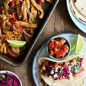 A top-down view of Sheet Pan Chicken Fajitas next to a plate with a stack of corn tortillas