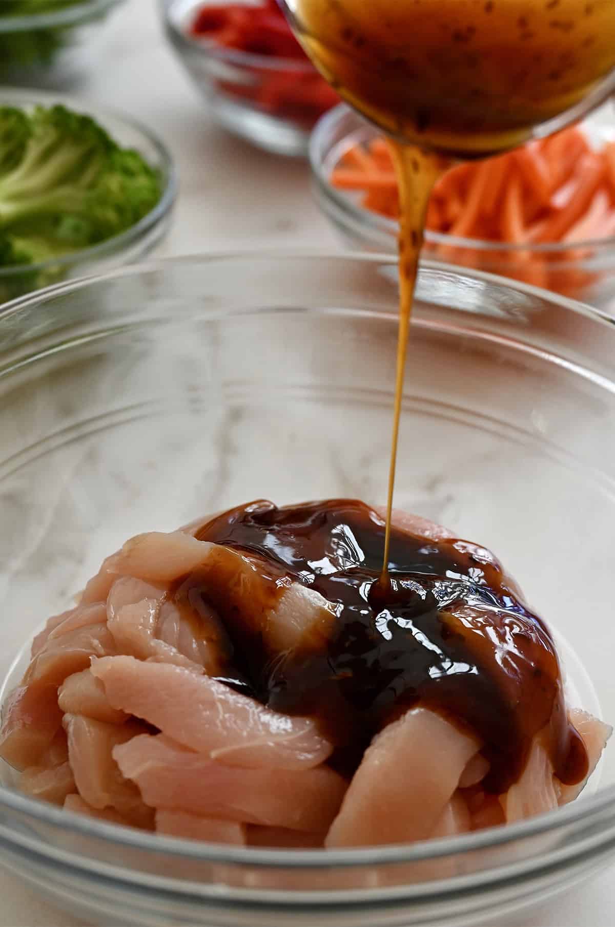 Pouring teriyaki sauce over strips of chicken breasts in a bowl.