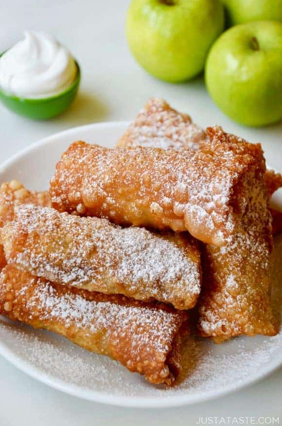 Apple pie egg rolls on a white plate with apples and whipped cream