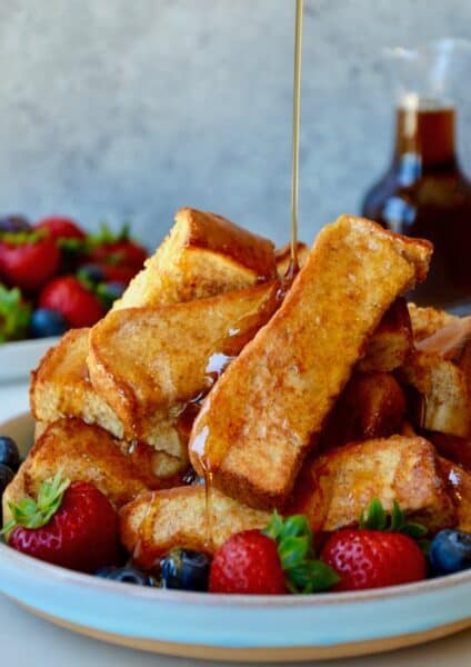 Baked French toast sticks pile on a plate with fresh berries with maple syrup being poured over them.