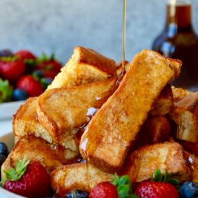 Baked French toast sticks on a plate with fruit and maple syrup being drizzled on top