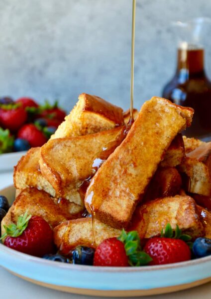 Baked French toast sticks on a plate with fruit and maple syrup being drizzled on top