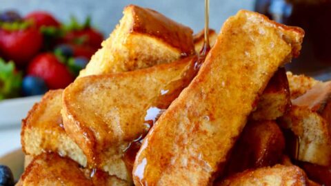 Baked French toast sticks on a plate with fruit and maple syrup being drizzled on top