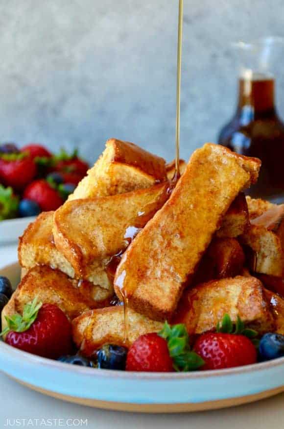 Baked French toast sticks on a plate with fruit and maple syrup being drizzled on top