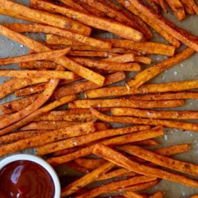 Baked sweet potato fries on a baking sheet with a bowl of ketchup