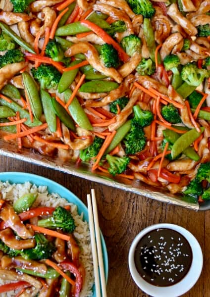 Sheet pan teriyaki chicken with broccoli, snap peas, red pepper and carrots on a rimmed baking sheet lined with foil. A plate with steamed rice topped with teriyaki chicken and chopsticks is nearby.