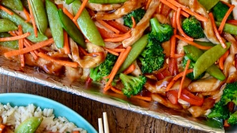 Sheet pan teriyaki chicken with broccoli, snap peas, red pepper and carrots on a rimmed baking sheet lined with foil. A plate with steamed rice topped with teriyaki chicken and chopsticks is nearby.