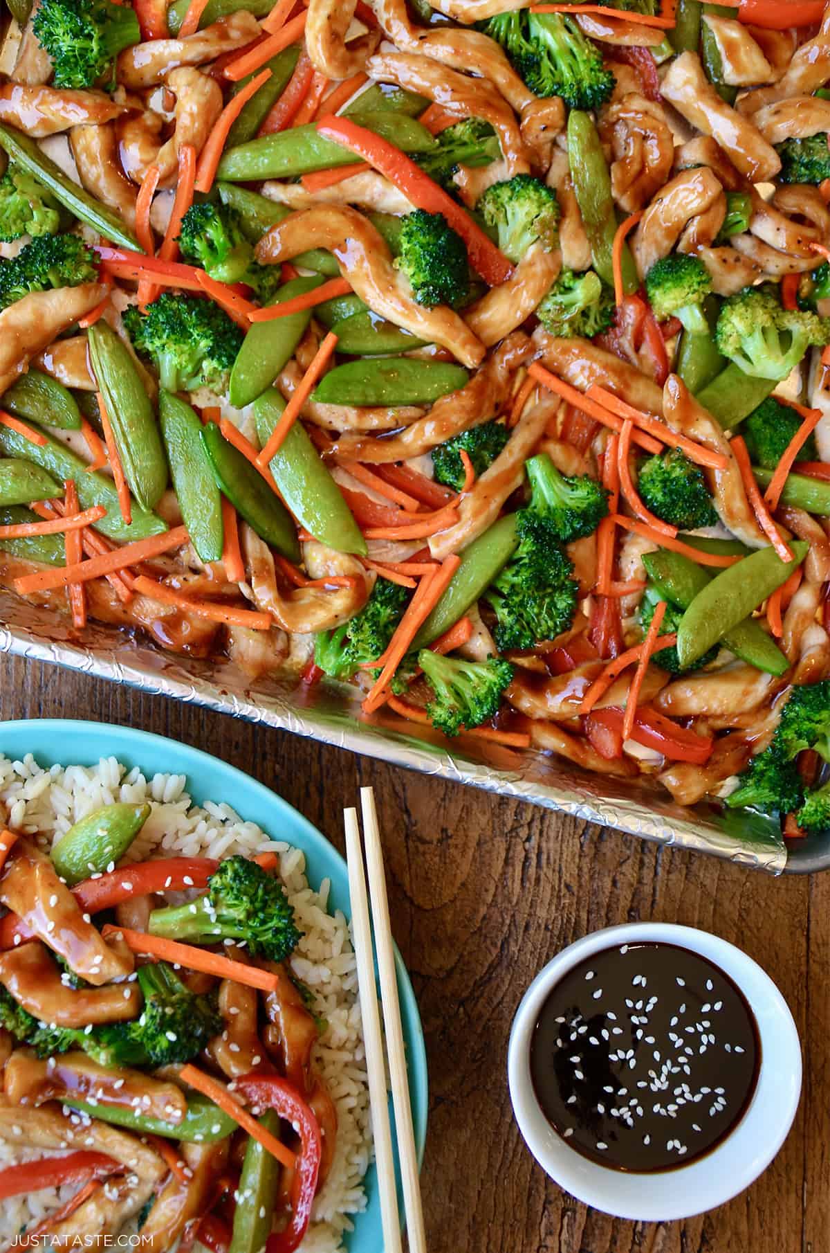 Sheet pan teriyaki chicken with broccoli, snap peas, red pepper and carrots on a rimmed baking sheet lined with foil. A plate with steamed rice topped with teriyaki chicken and chopsticks is nearby.