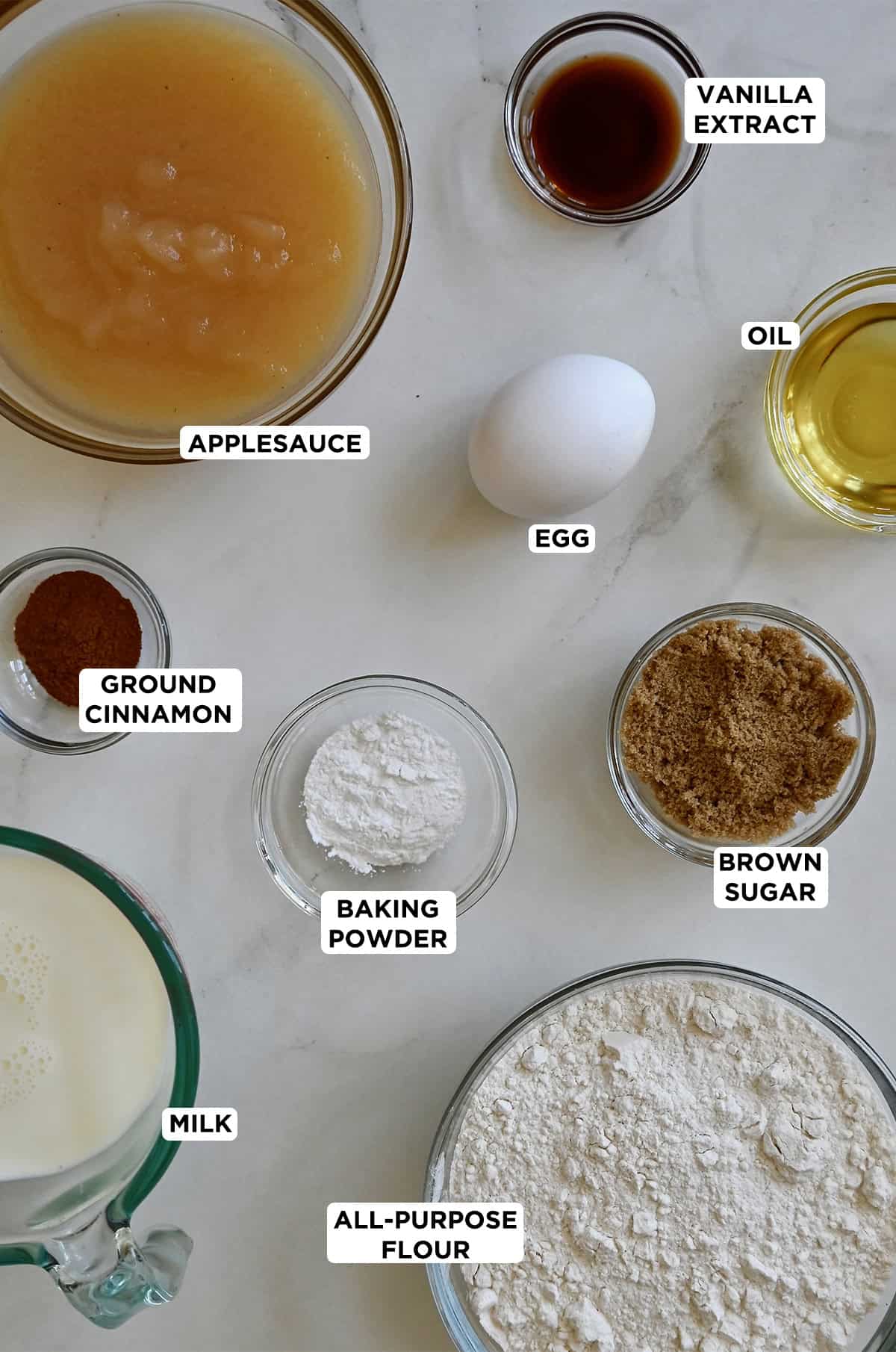 Various sizes of glass bowls containing the ingredients to make apple pancakes, including applesauce, flour, ground cinnamon, brown sugar, an egg, oil, baking powder and milk.