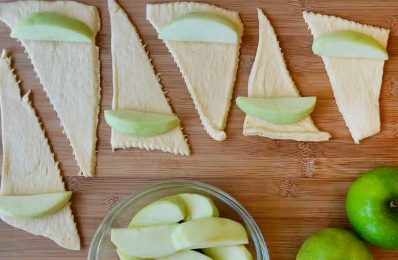 Uncooked crescent rolls and apple slices being rolled up on a cutting board