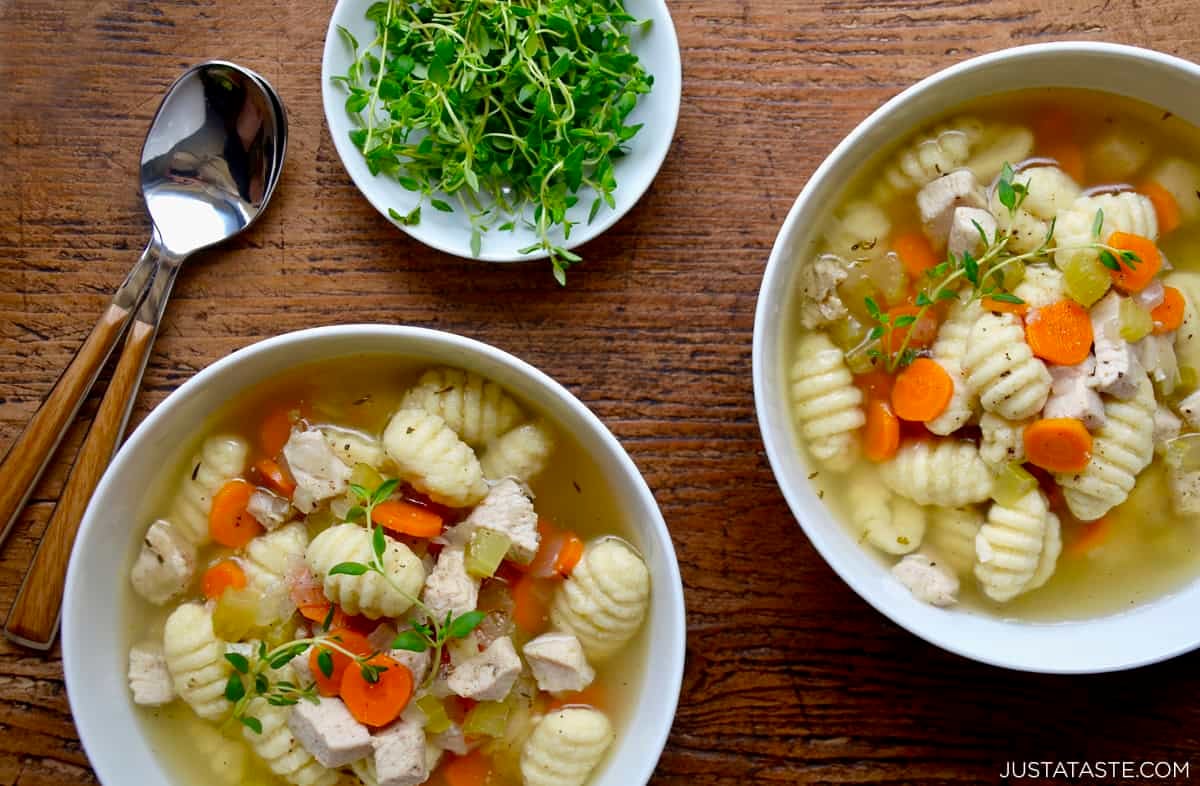 Two bowls containing homemade chicken soup with gnocchi, carrots and celery topped with a sprig of fresh thyme. Soup spoons and a bowl filled with fresh thyme is nearby.