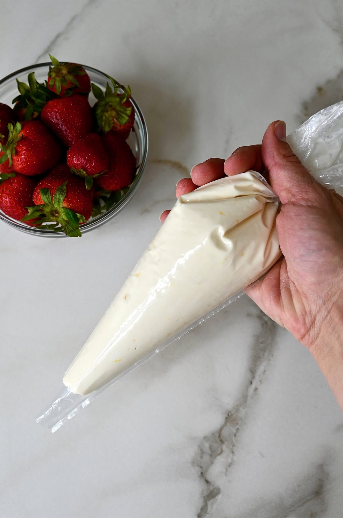 A hand holds a plastic piping bag containing lemony cream cheese filling for Danish pastries. A bowl filled with fresh strawberries is nearby.