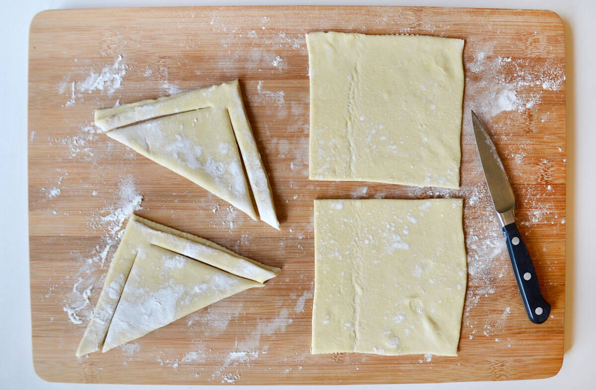 Two triangle-shaped puff pastry dough with slits next to two squares of puff pastry dough on a cutting board with a sharp knife.