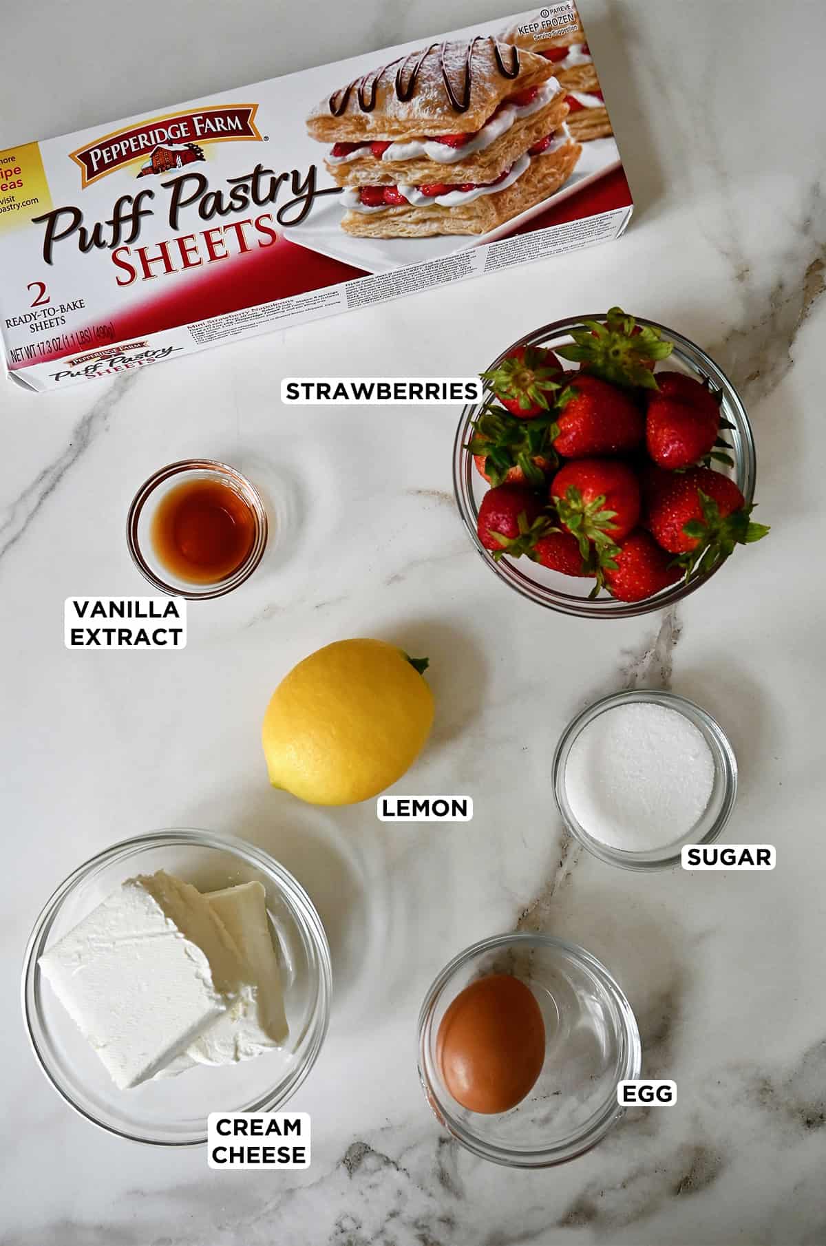 A box of puff pastry next to a lemon and various sizes of glass bowls containing vanilla extract, strawberries, cream cheese and an egg.