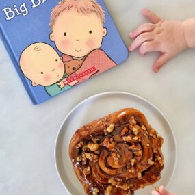 A baby's hands reaching for a sticky bun and a book
