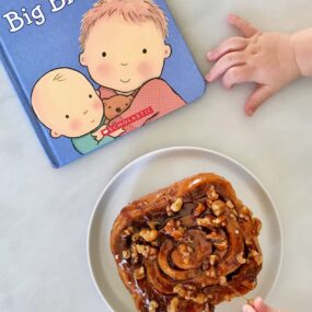 A baby's hands reaching for a sticky bun and a book