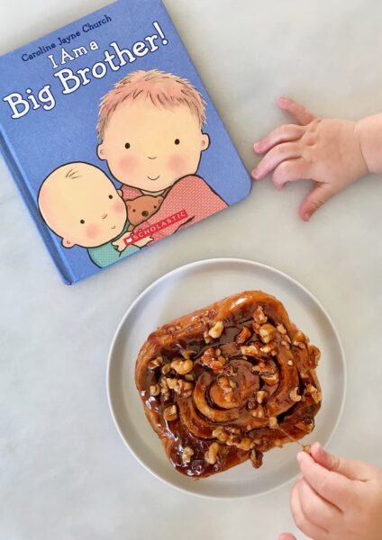 A baby's hands reaching for a sticky bun and a book