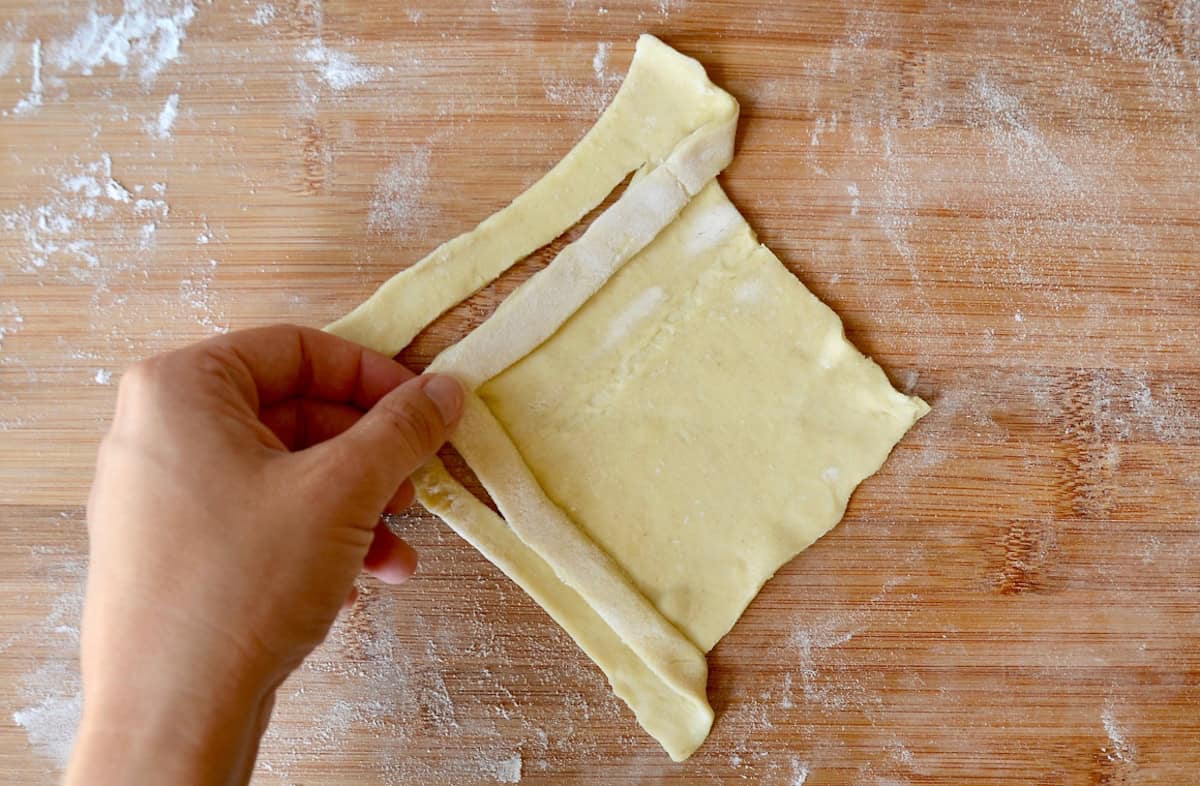 A hand folds puff pastry to form a diamond shape.