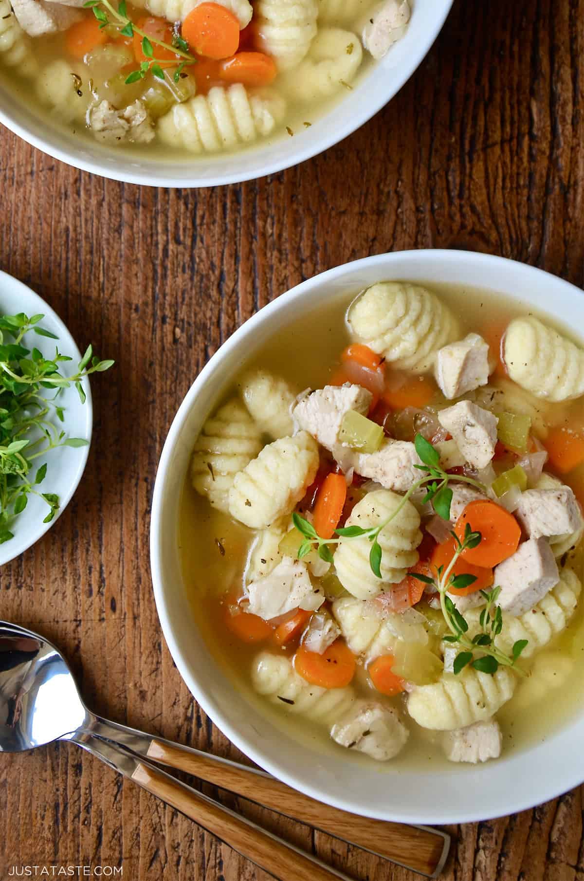 Homemade chicken soup with gnocchi, carrots and celery topped with a sprig of fresh thyme. Soup spoons and a bowl filled with fresh thyme is nearby.